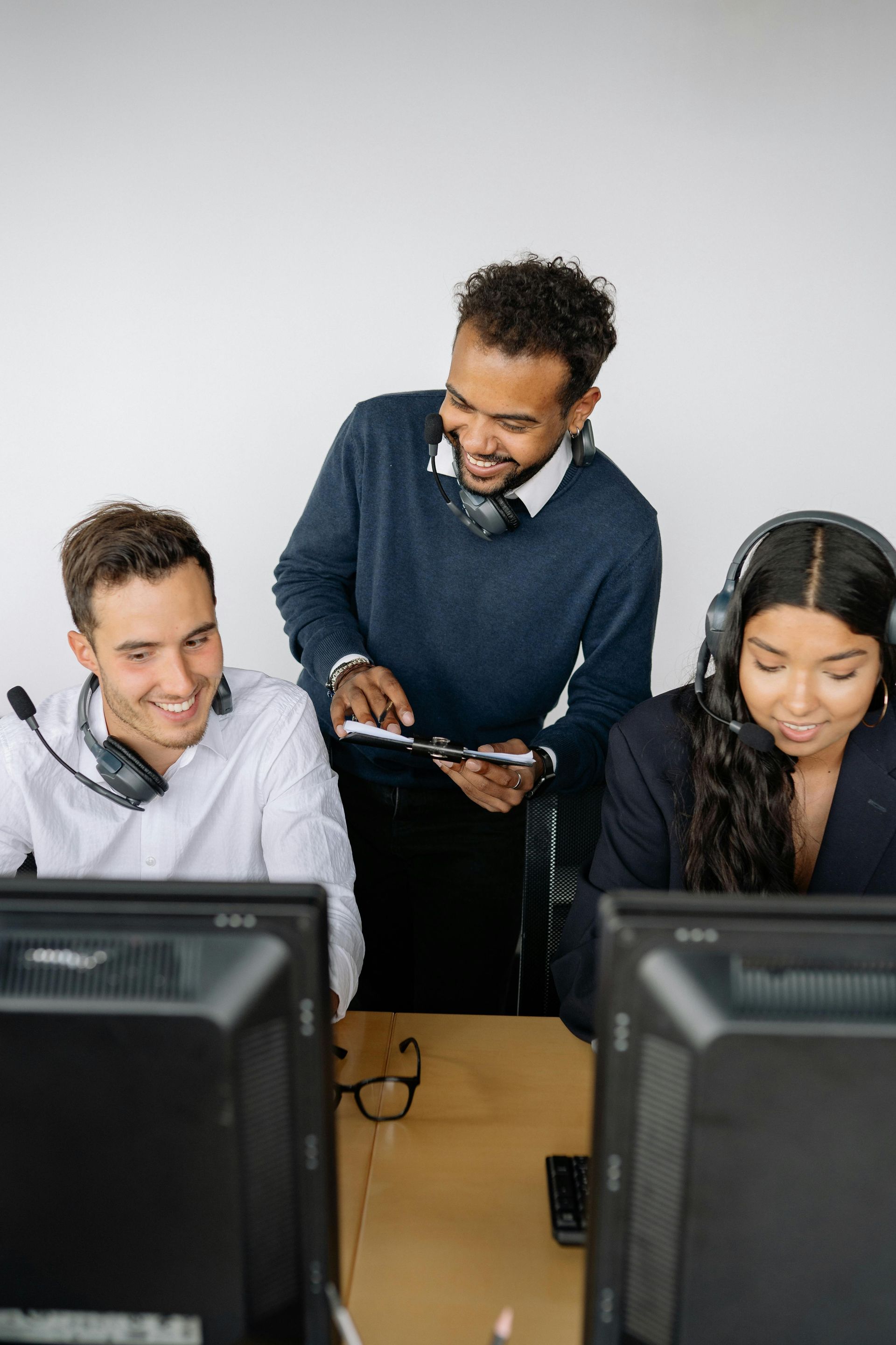 Three people in a call center; a supervisor points to a tablet, smiling, while employees smile at their computers.