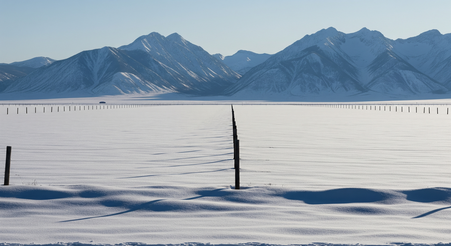 Snow-covered field with a fence in front of snow-capped mountains under a clear blue sky.