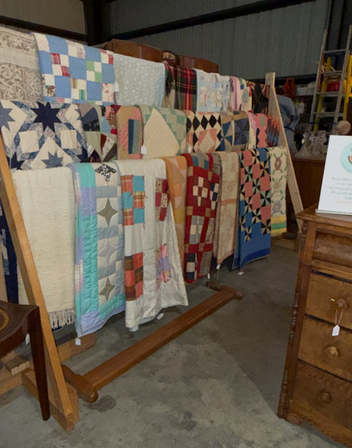 A wooden display rack holds a variety of colorful patterned quilts in a rustic shop setting.