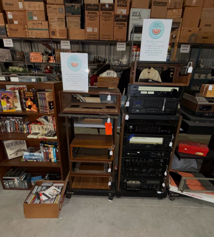 A wooden shelf, a media stand, and a stack of audio equipment in a storage room with cardboard boxes on shelves.
