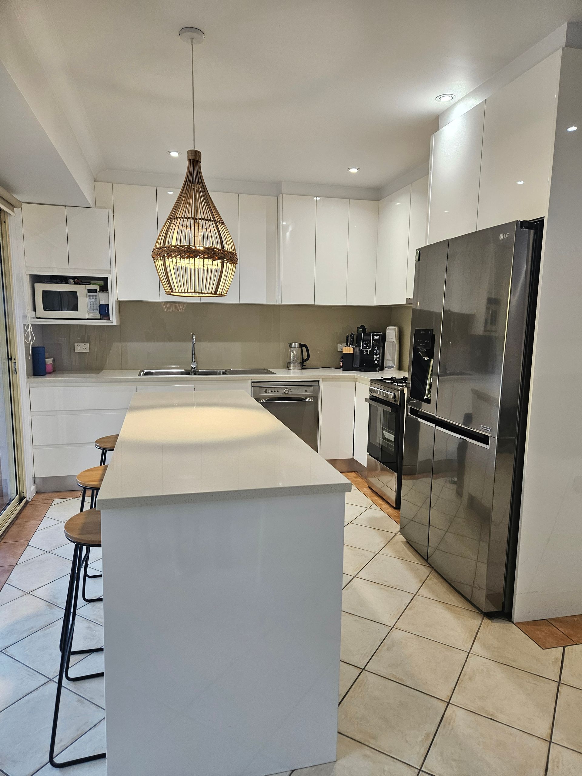 Bright white kitchen with island, woven pendant light, tiled floor, and black refrigerator.