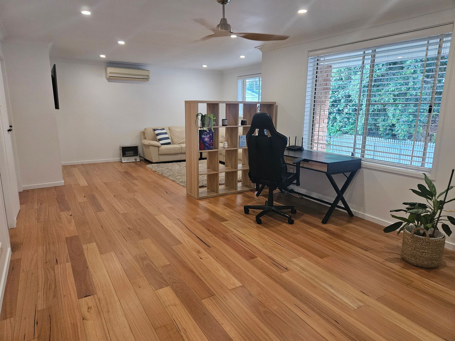 Spacious living room with hardwood floors, a desk by large windows, sofa, and potted plants.
