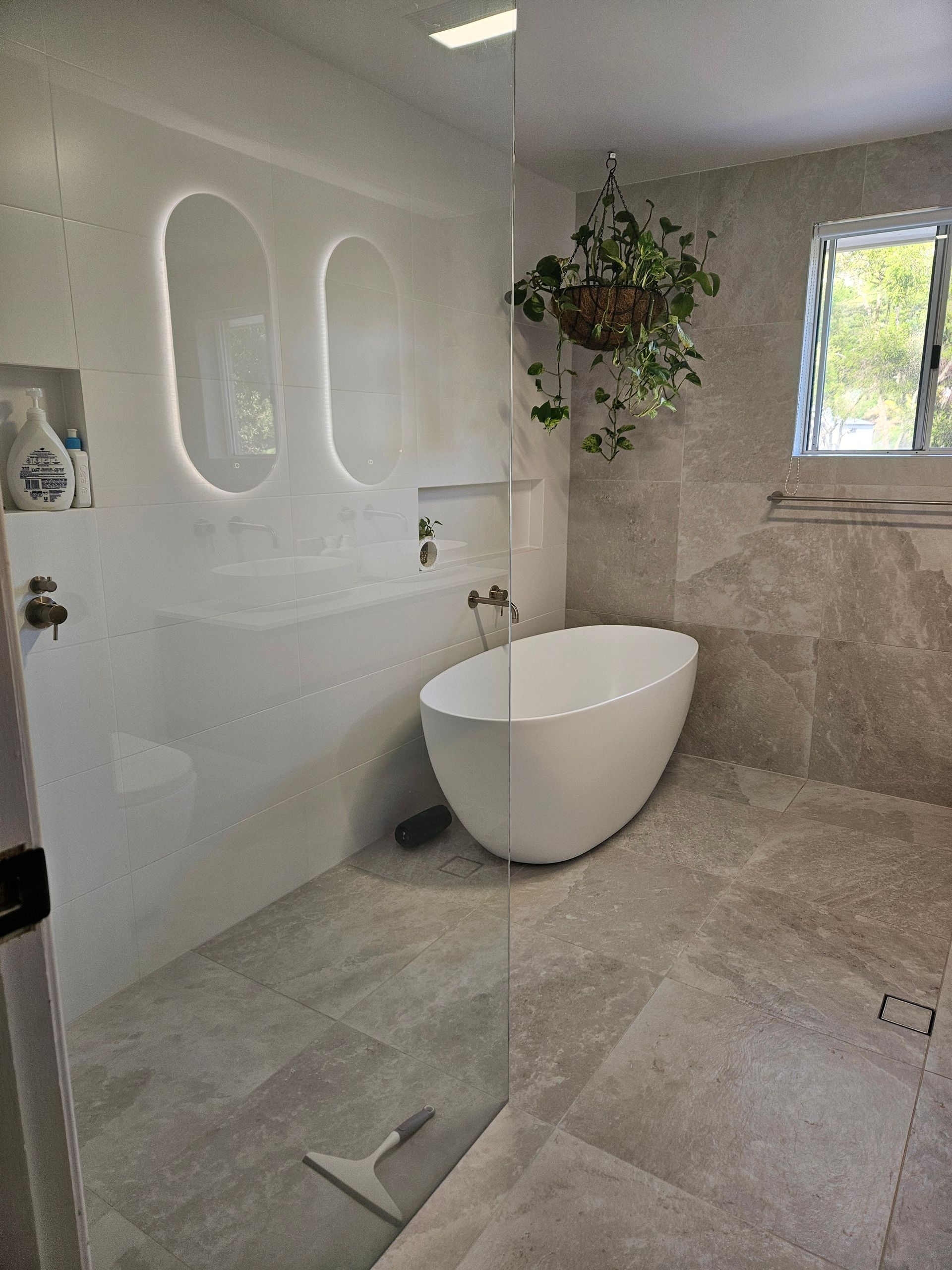 Modern bathroom with a freestanding tub, large shower, beige tile, and a hanging plant.