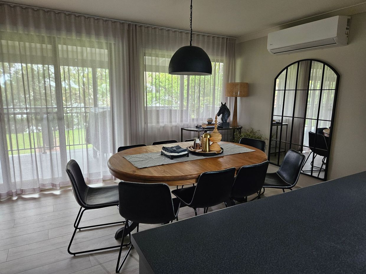 Dining room with round table, black chairs, hanging pendant light, large windows, and a wall mirror.