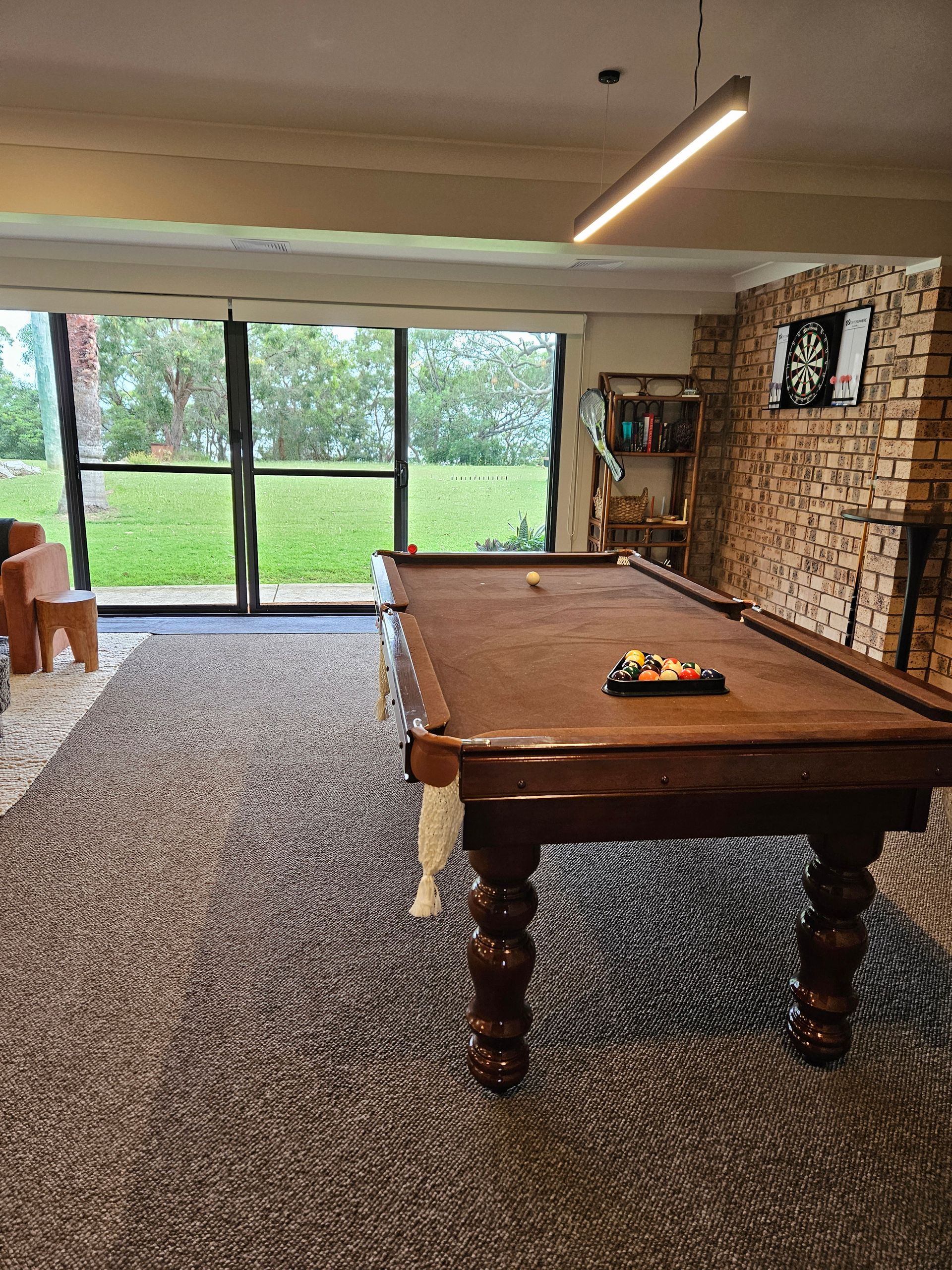 Spacious lounge with a pool table, patterned carpet, brick wall, and large glass doors overlooking a green yard