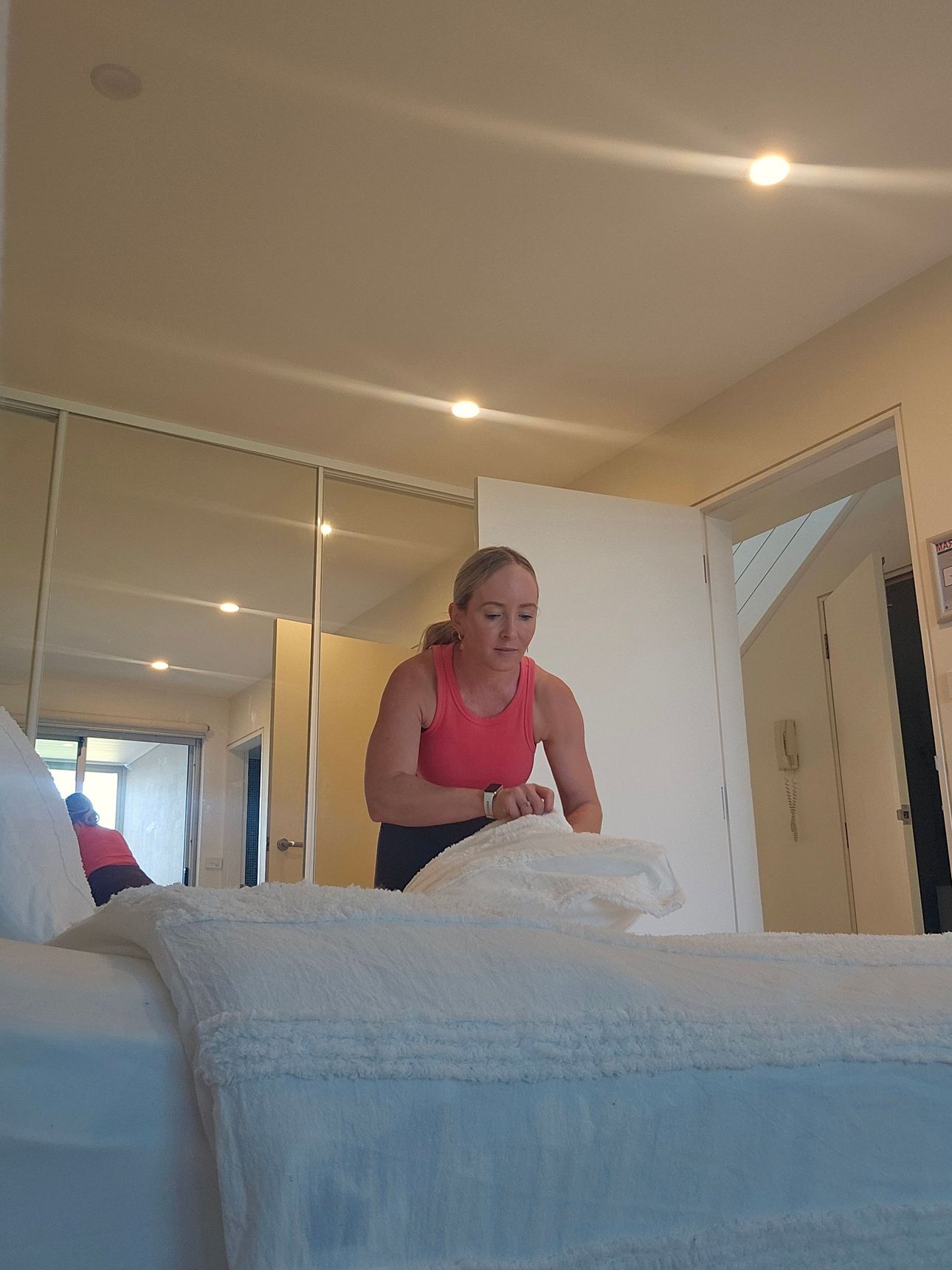 Person in a pink top making a bed in a bright bedroom with mirrored closet doors.