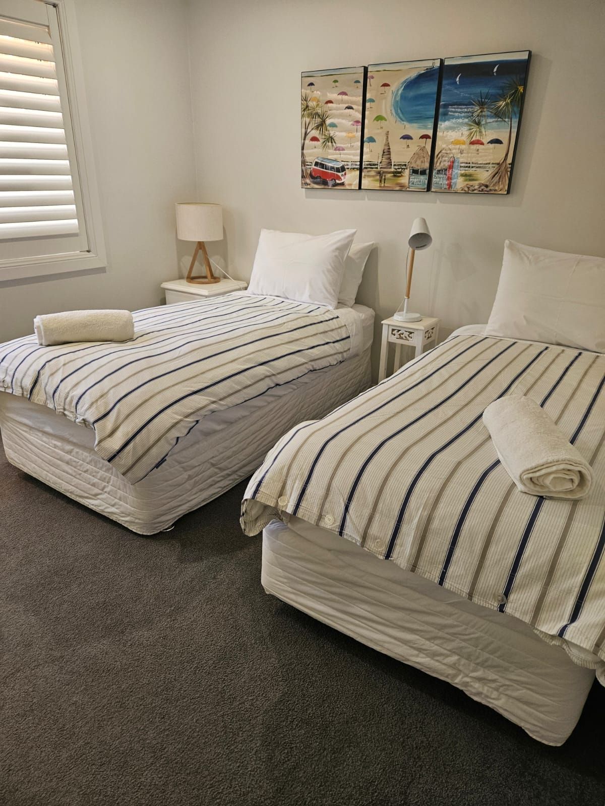 Twin beds with white striped bedding in a bright bedroom, with a bedside lamp and beach wall art.