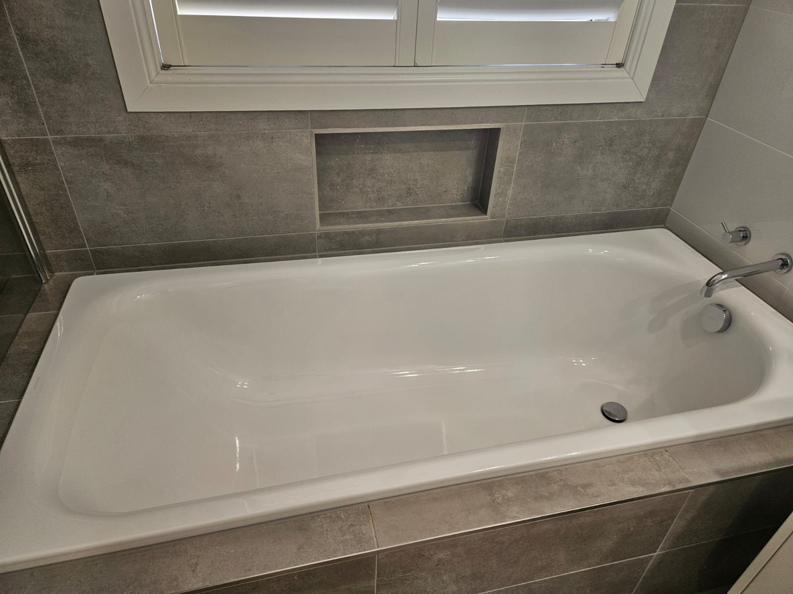 White bathtub with tiled surround beneath a window in a bathroom.