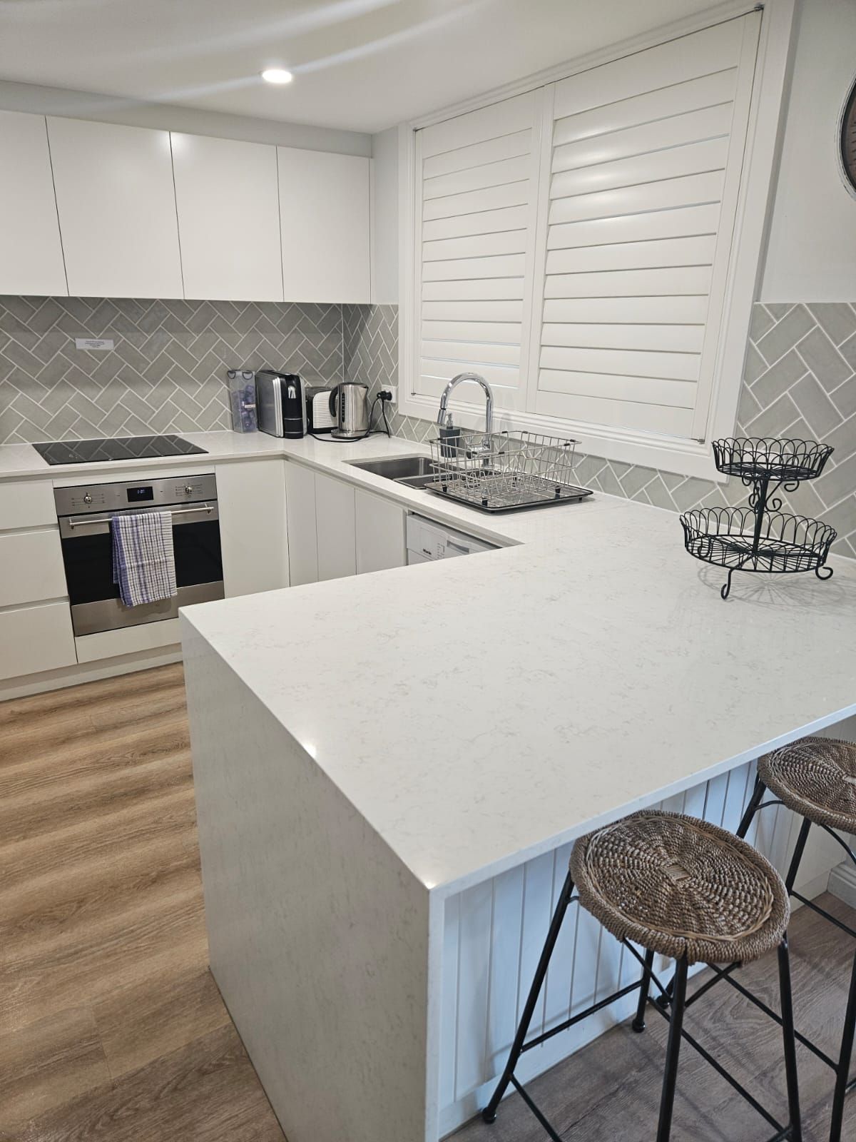 Bright modern white kitchen with island, barstools, stainless appliances, and wood-look flooring.