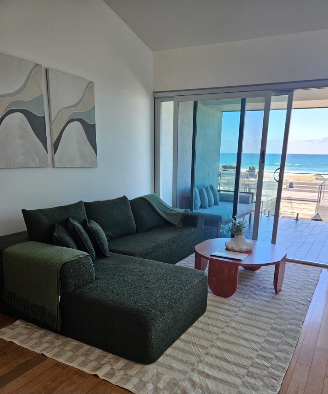 Modern living room with dark green sectional, red coffee table, and ocean view through sliding glass doors