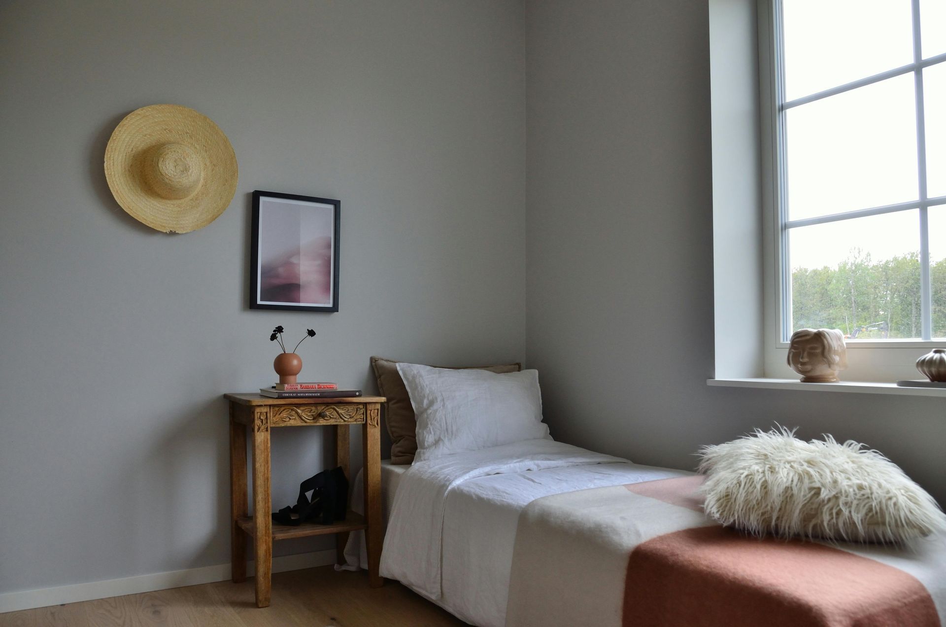 Minimal bedroom with a single bed, wooden table, framed art, straw hat, and a window letting in daylight