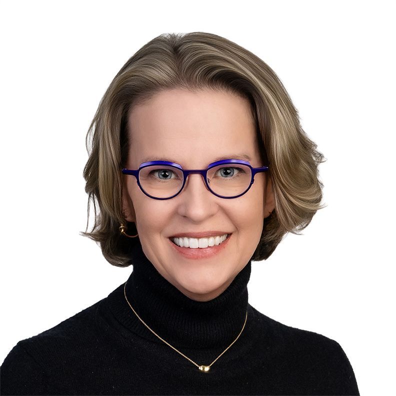 Headshot of a smiling person wearing pink glasses, a black blazer, and white top on a white background