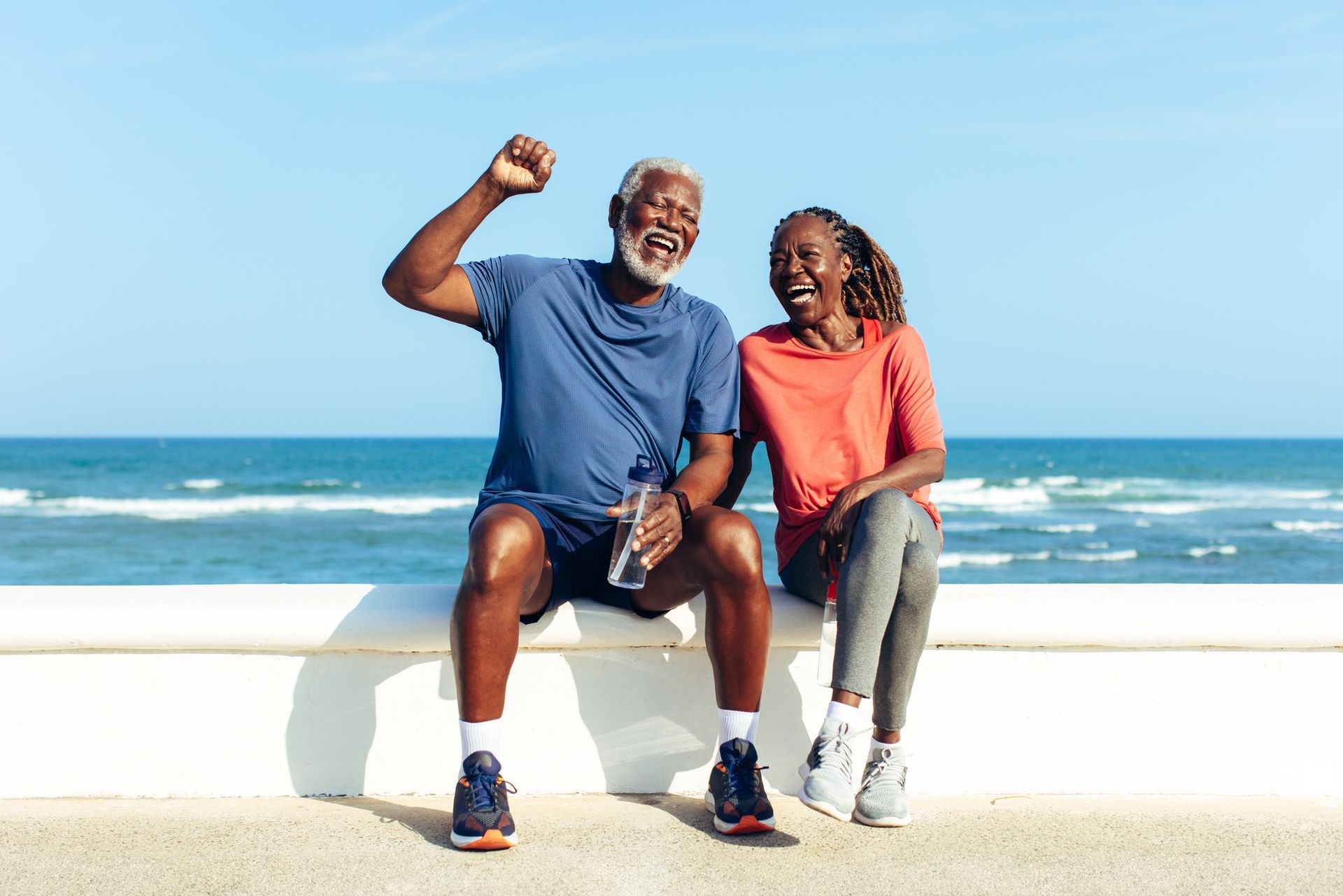 Two people sitting on a seaside wall, smiling and waving, with ocean waves in the background.