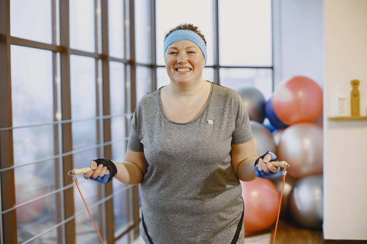 Woman in a gym smiling while holding jump ropes, with exercise balls in the background.