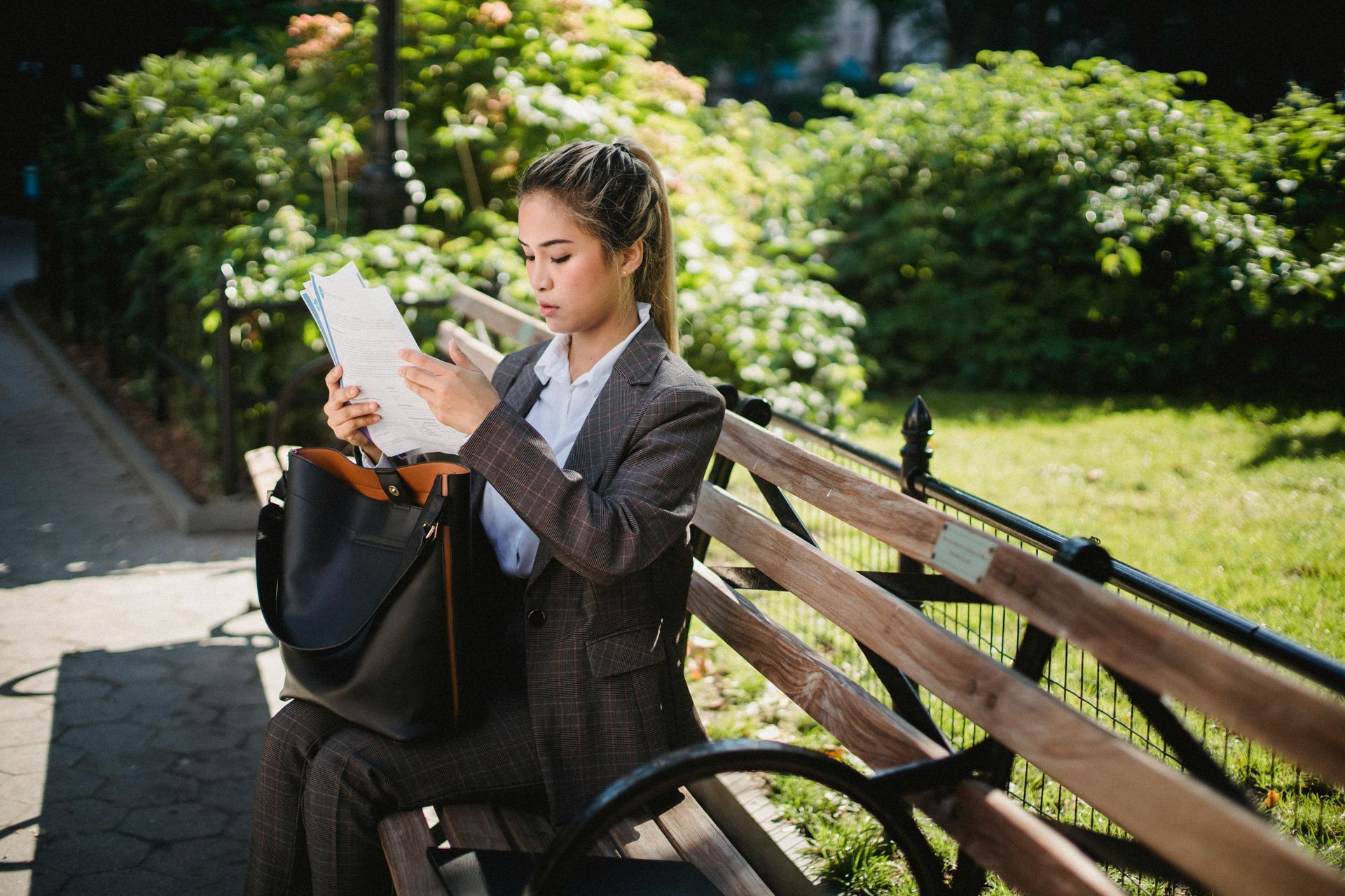 Person reading a paper on a park bench beside a black bag in a sunny green park