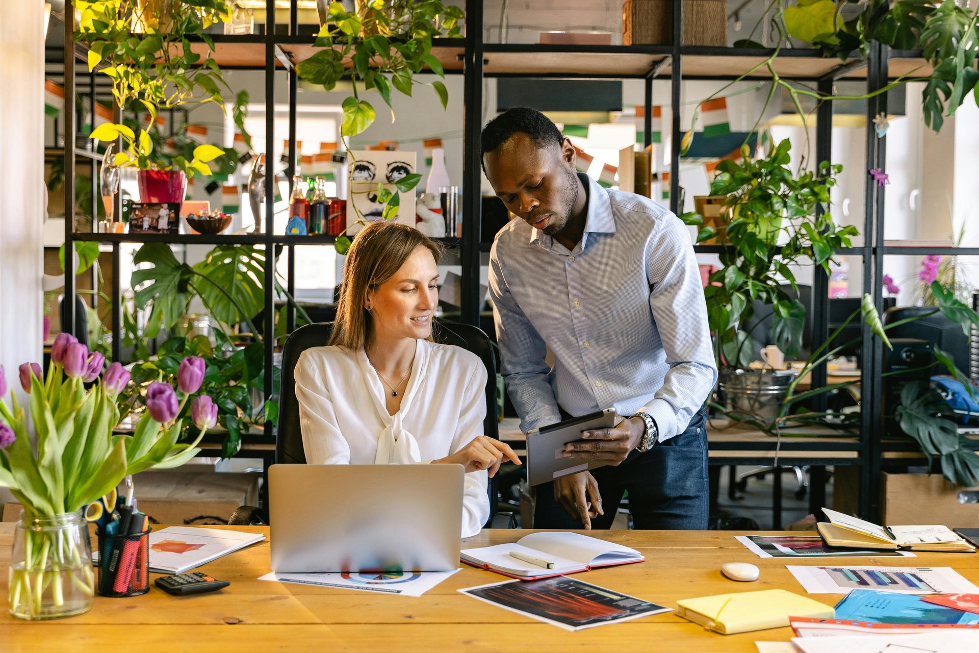 Two coworkers reviewing documents at a desk in a bright office with plants and a laptop