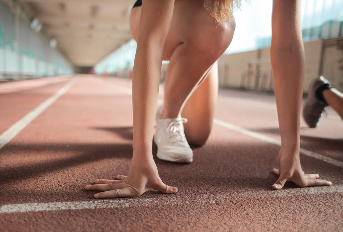 Runners crouched at a track starting line, with hands on the red track surface and white shoes visible.