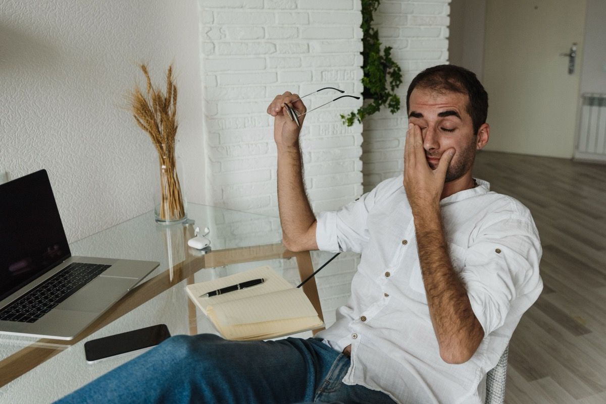 Man seated at a desk with laptop and notebook, rubbing his face in a bright room