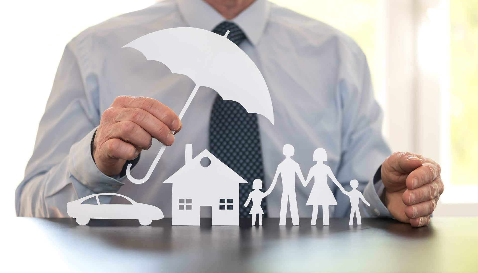 Businessman holding an umbrella over paper family, house, and car icons, symbolizing insurance protection