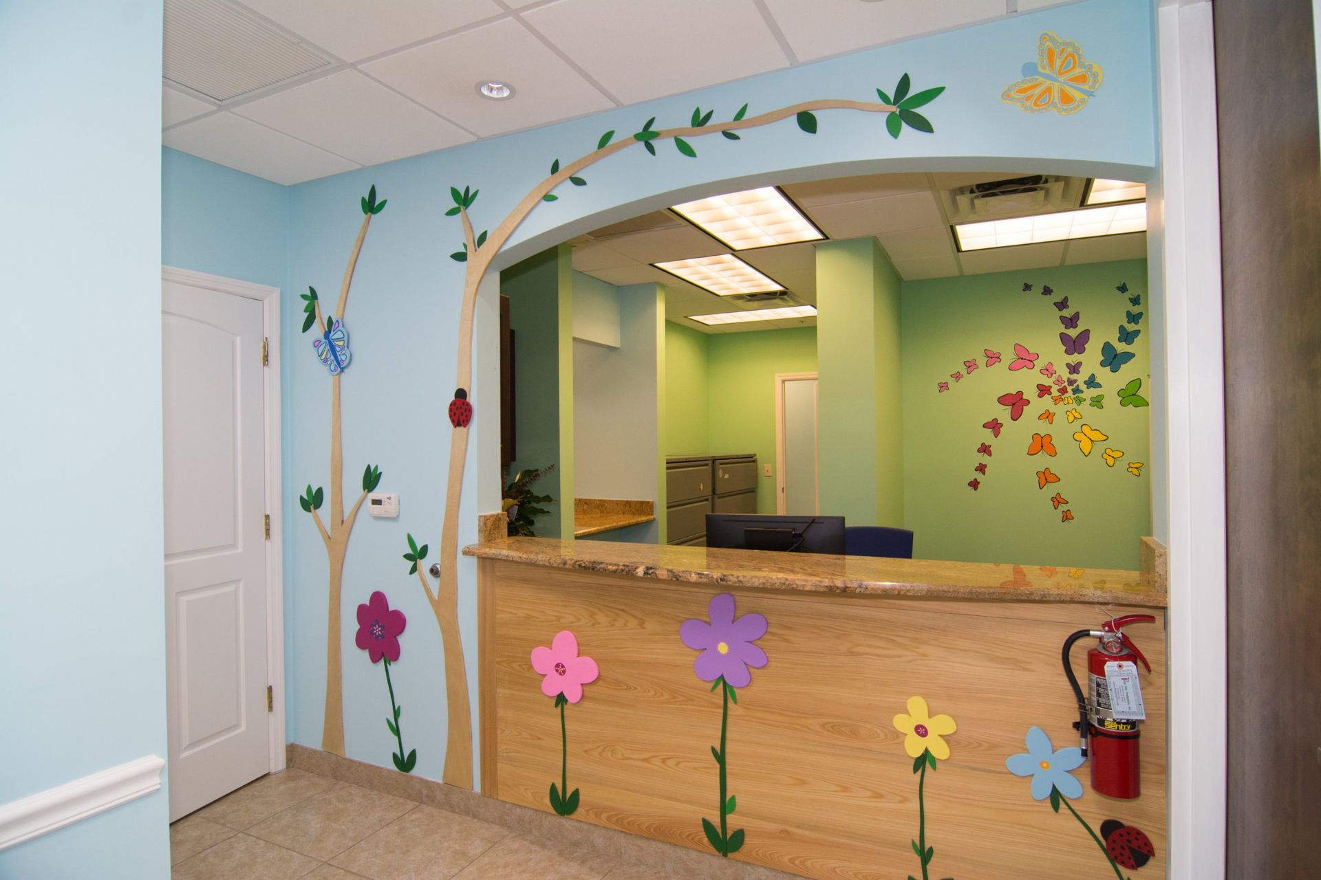 Light Blue wall with flower decals and butterflies, wooden counter with nobody behind it, white door on the right.