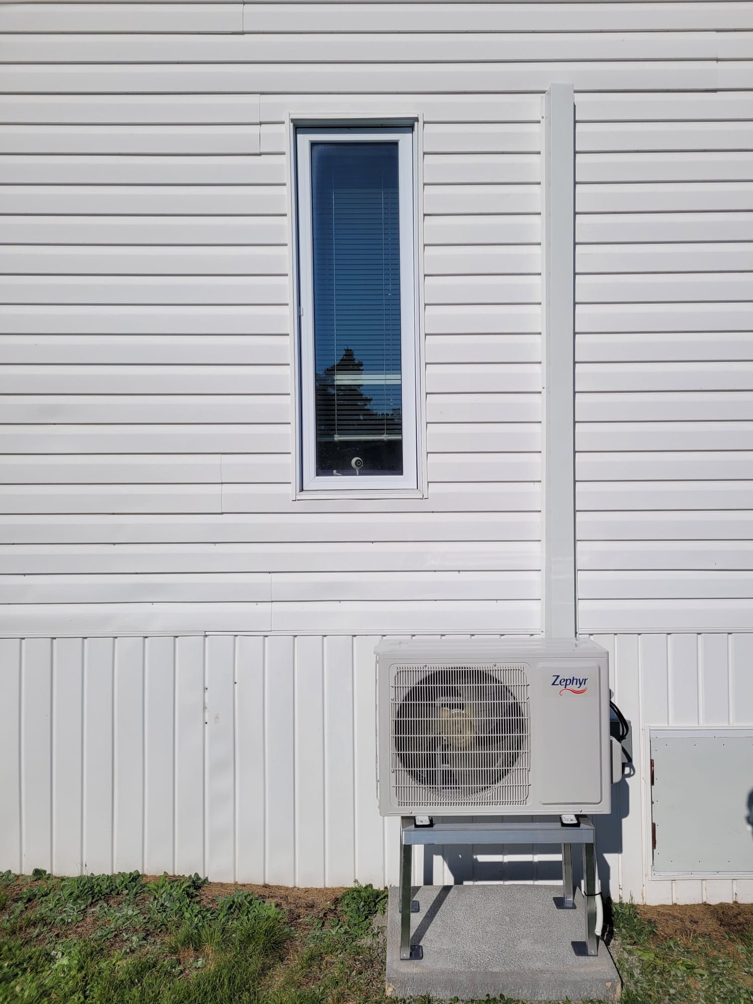 Revêtement extérieur blanc pour maison, avec une fenêtre étroite et une pompe à chaleur montée à l'extérieur sur un support.
