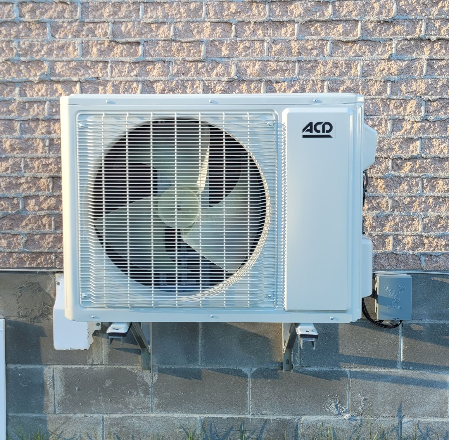 Unité de climatisation extérieure blanche fixée sur un mur de briques, avec ventilateur et tuyaux apparents.