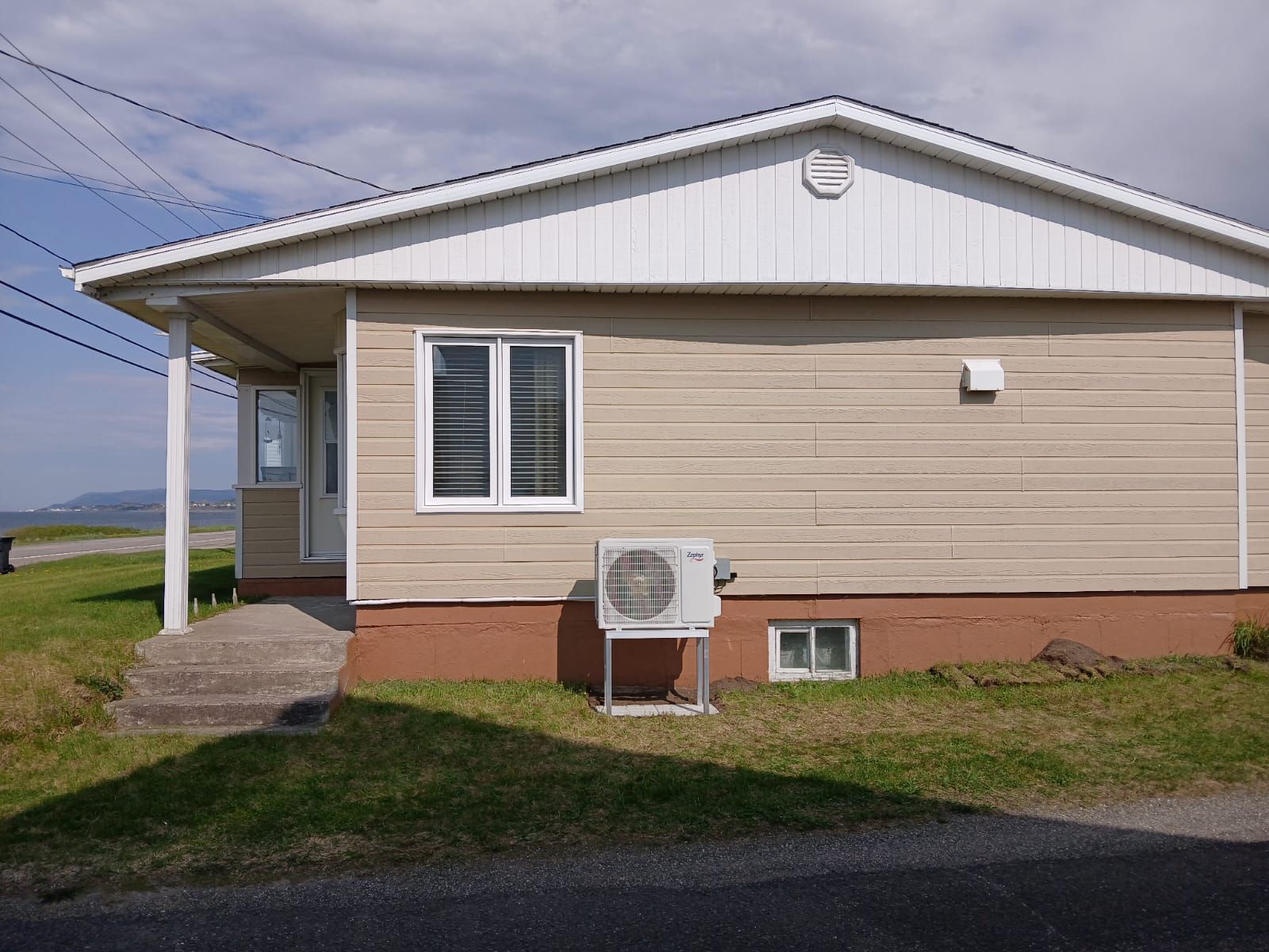 Petite maison beige avec des boiseries blanches, un porche et un climatiseur, sur un terrain gazonné.