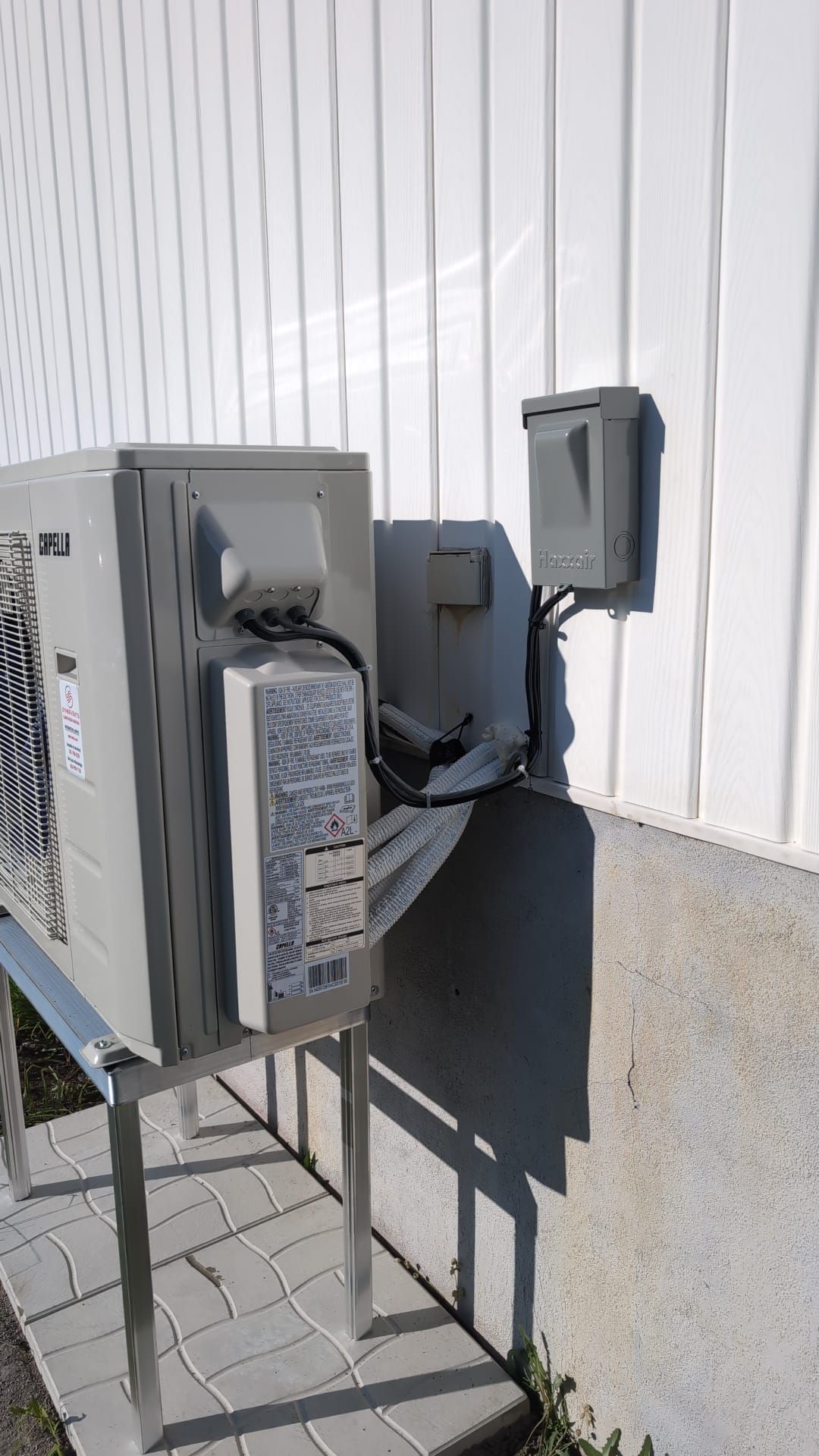 Unités électriques et de CVC extérieures montées contre un mur blanc, avec câbles et conduits visibles.