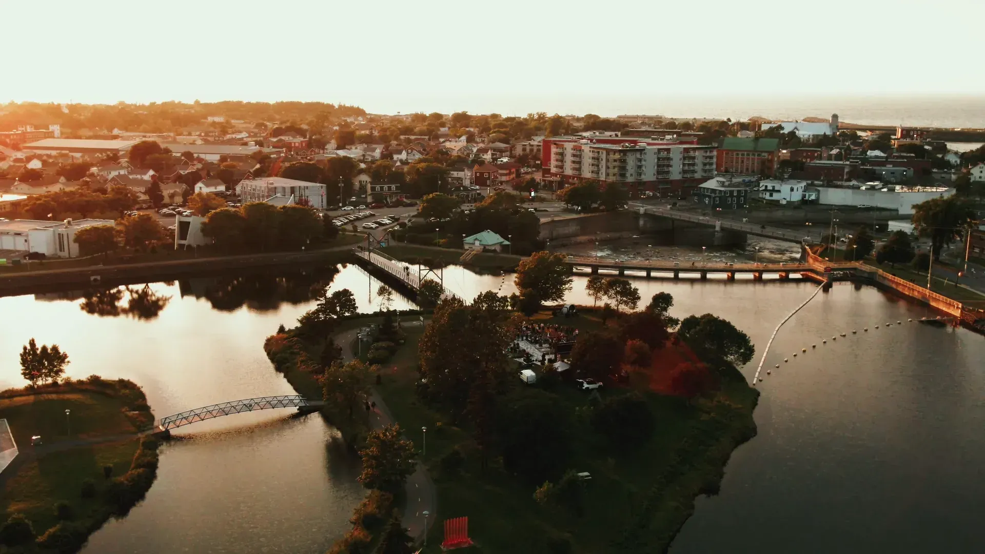 Vue aérienne au coucher du soleil d'une rivière, d'un pont et de bâtiments urbains baignés d'une chaude lumière orangée.