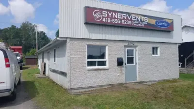 Petit bâtiment commercial blanc avec une enseigne Synerventes et des véhicules garés à l'extérieur.