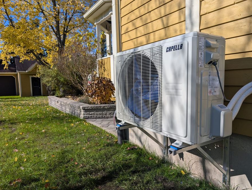 Unité de climatisation extérieure à côté d'une maison jaune, avec une cour gazonnée et des arbres d'automne à proximité