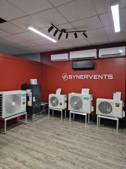 Showroom Synervents avec cinq ventilateurs industriels sur pieds contre un mur rouge et des luminaires au plafond carrelés