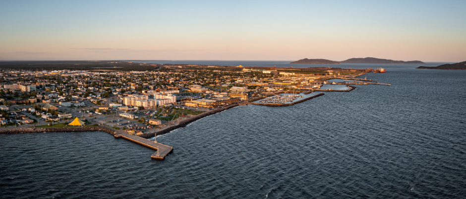 Vue aérienne d'une ville côtière au coucher du soleil, avec un long port et des eaux bleues calmes.