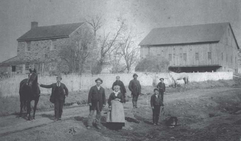 Six people and a horse stand in front of two barns in a rural yard.