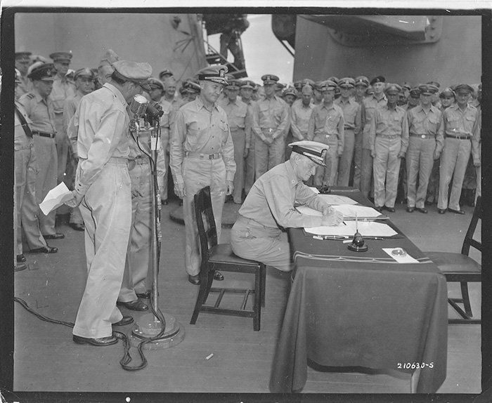 Military officer signs document at table while uniformed soldiers stand at attention behind him.
