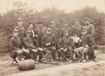 Group portrait of uniformed men posing outdoors with a cannon in front, in a sepia-toned historical photo