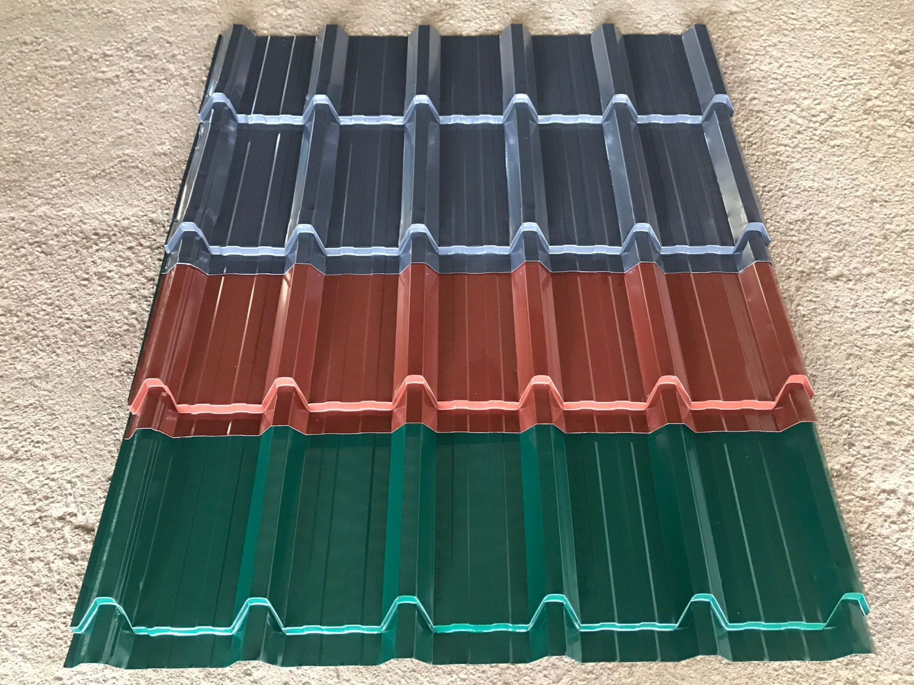 Tres paneles de techo de metal corrugado: gris, rojo y verde, apilados sobre una superficie marrón.