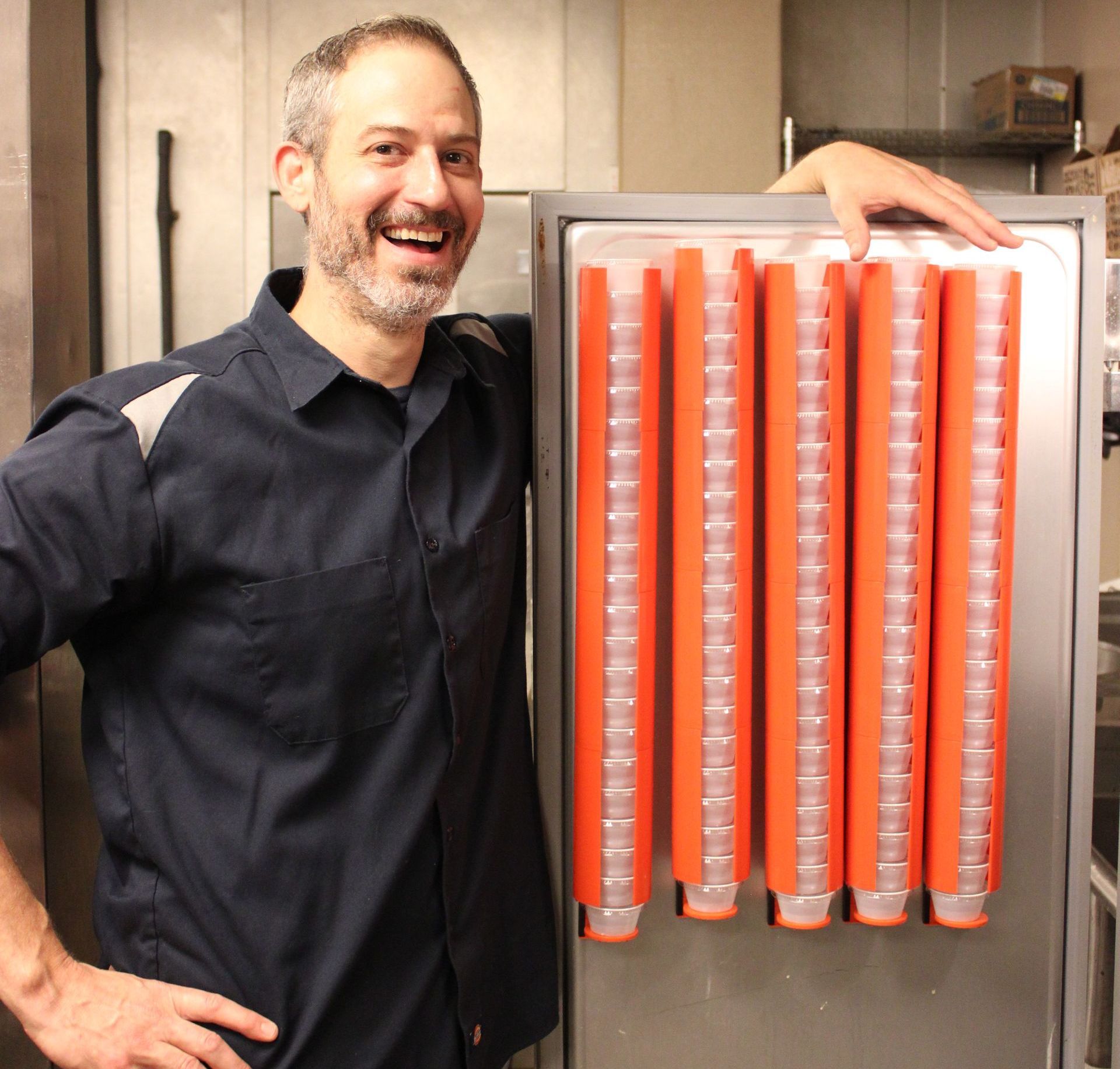 Man smiles beside a mounted ramekin dispenser with orange tubes; inside a commercial kitchen.