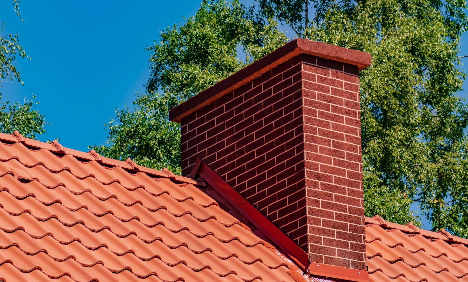 Red brick chimney on a terracotta-tiled roof against blue sky and green trees