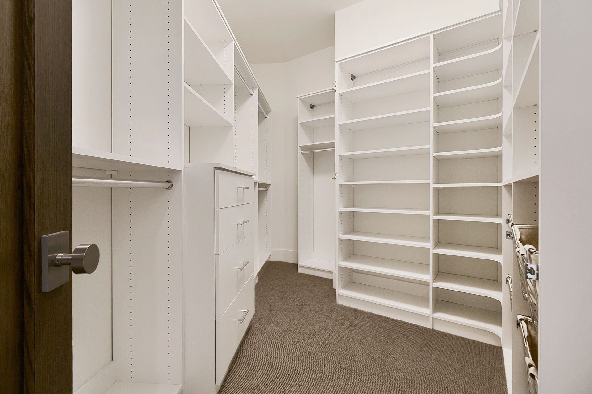 Empty white walk-in closet with shelves, drawers, and a hanging rod; beige carpet.