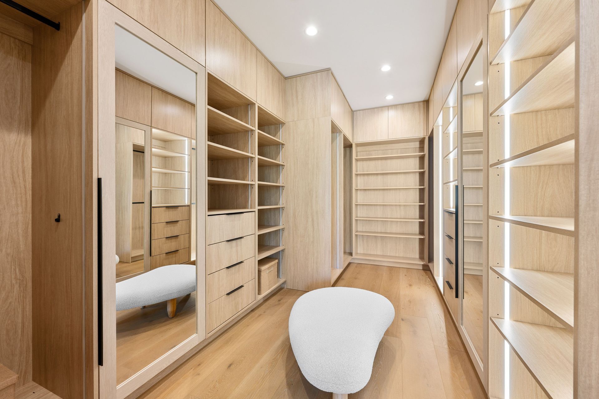 Wooden walk-in closet with shelves, drawers, and a large mirror, featuring a white ottoman.