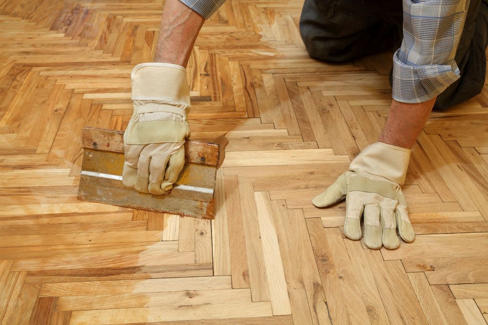 A Man is Painting a Wooden Floor With a Spatula — Norwood Floor Sanding in Byron Bay, NSW