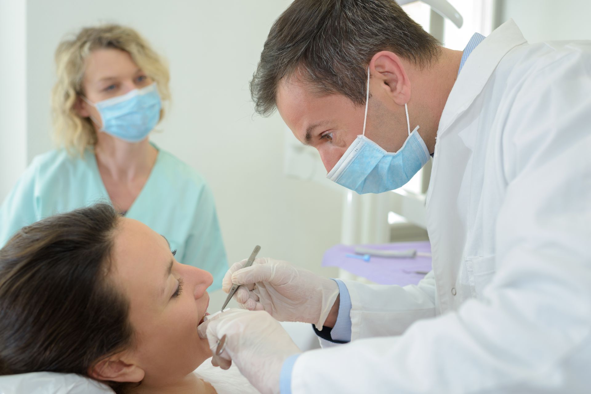A dentist is inspecting the teeth of a patient. A dental assistant is in the background. A dentist is inspecting the teeth of a patient. A dental assistant is in the background.