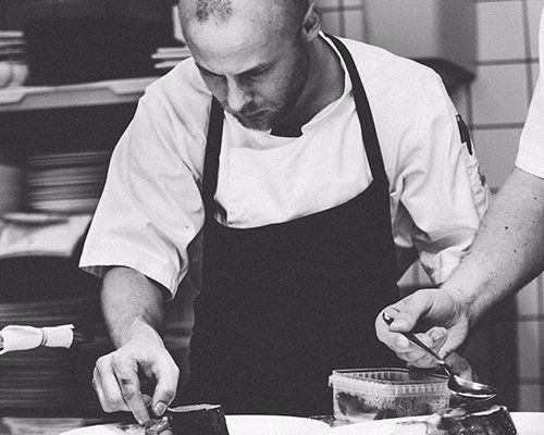 Chef in white apron plating food in a kitchen, focused on arranging a dish.