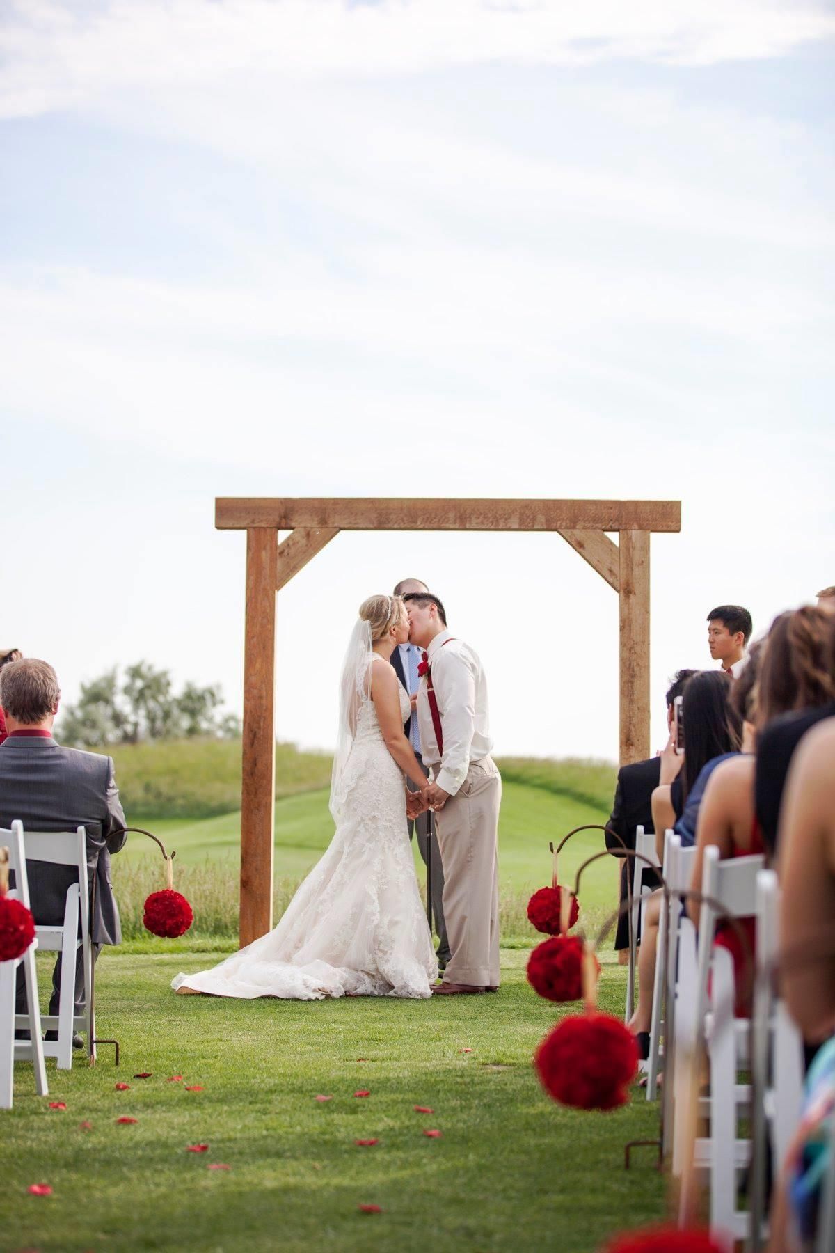 A bride and groom are kissing at their wedding ceremony under a wooden arch.