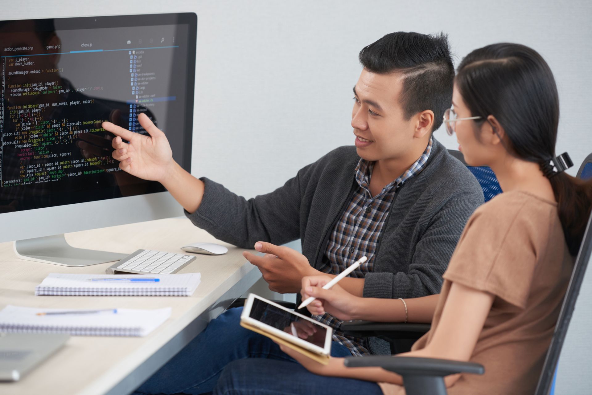 Man points at code on a monitor, explaining to a woman holding a tablet in an office setting.