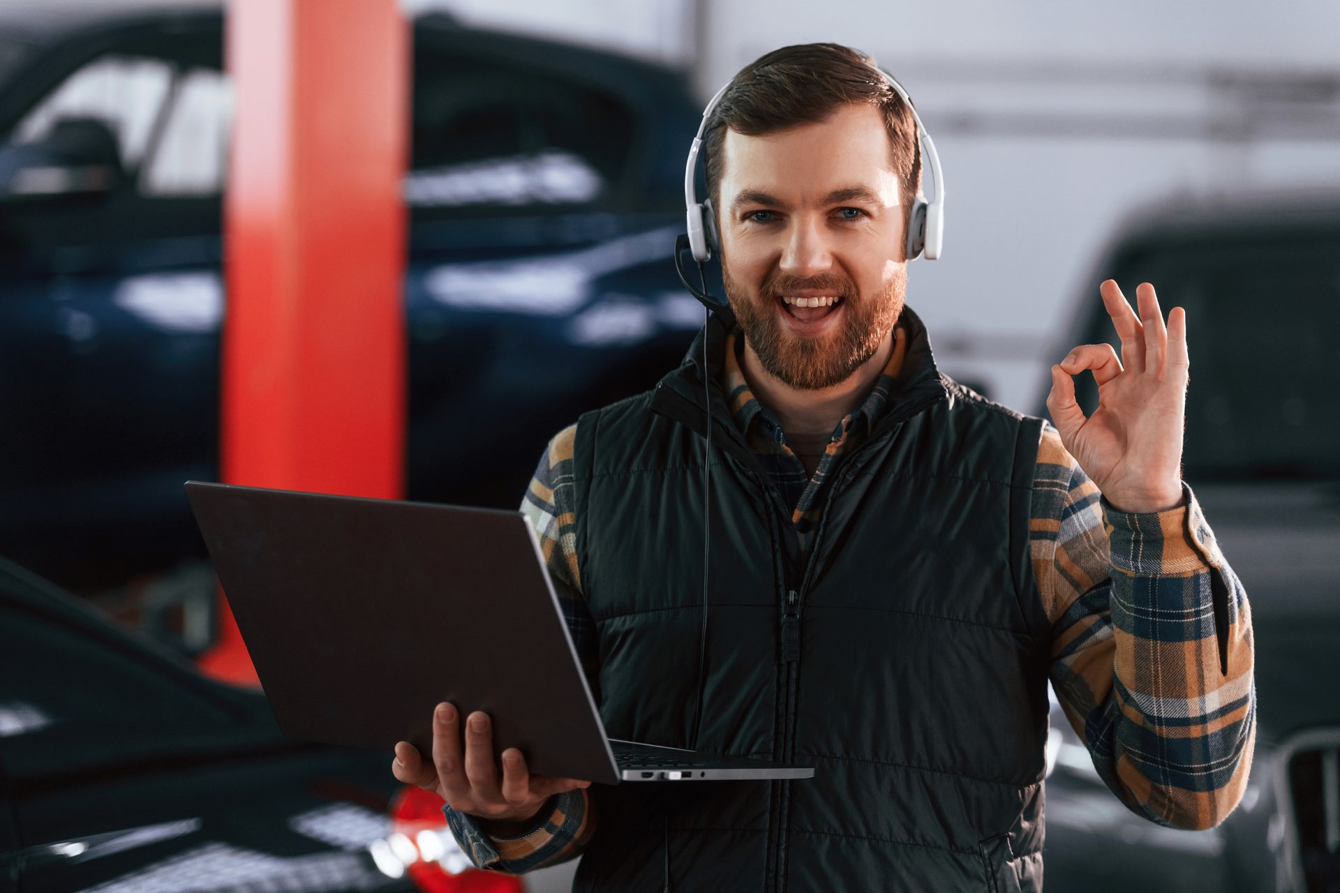 Mechanic in a garage holding a laptop, wearing a headset, and giving the