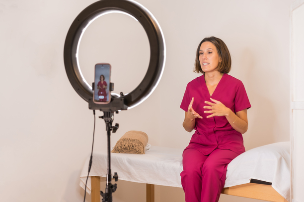Woman in pink scrubs sits on a massage table, gesturing while filming with a phone and ring light.