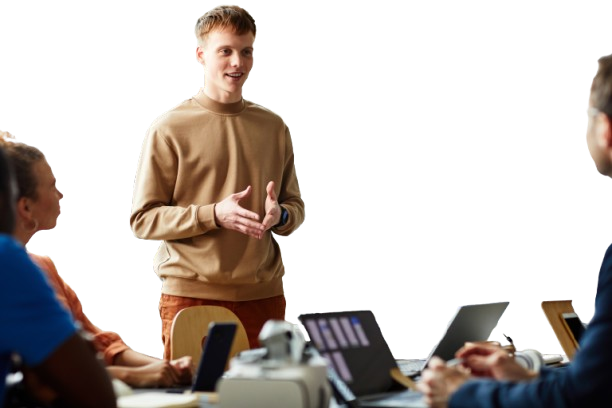 Man in brown sweater presents to a seated group with laptops at a table, gesturing with hands.