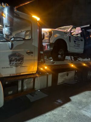 A white flatbed tow truck at night, transporting a white work truck on its bed.